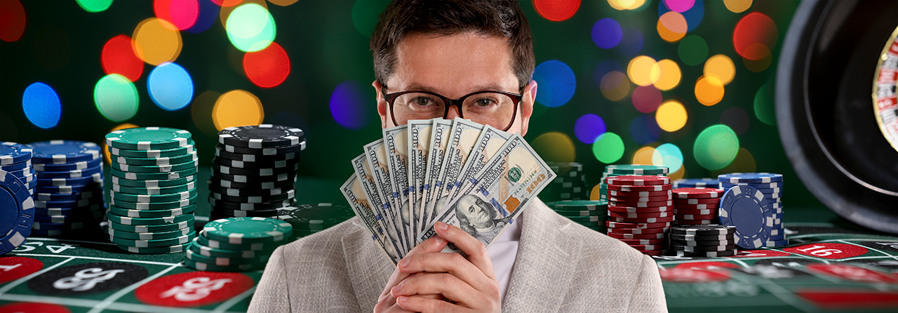 A smiling man fanning out a handful of hundred-dollar bills in front of stacks of casino chips on a roulette table