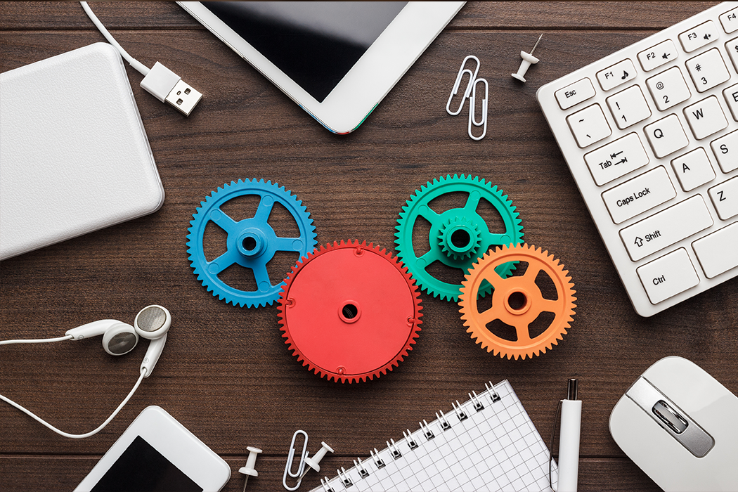 Four colorful interlocking gears on a desk, surrounded by a keyboard, mouse, tablet, phone and notepad