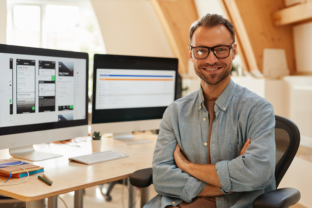 A smiling man with his arms crossed, sitting at a desk in front of two computer screens